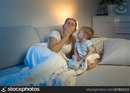 Adorable baby boy sitting on bed and looking at yawning tired mother