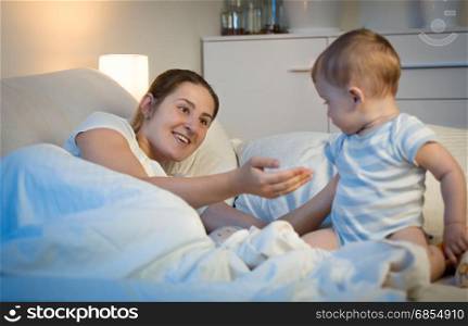 Adorable baby boy sitting on bed at night and looking at mother