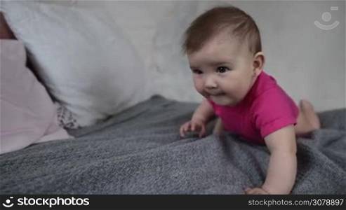 Adorable baby girl playing on bed, making her first step, learning to crawl. Portrait of happy infant child having fun on bed while family relaxing at home. Slow motion.