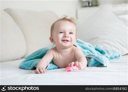 Adorable baby lying under blue towel on bed and holding plastic toy