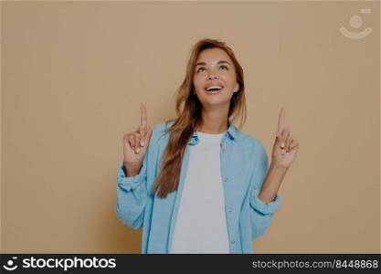 Adorable brunette female with long straight hair wears casual white tshirt and blue shirt, indicates upwards on blank copy space, notices something on ceiling, stands against beige wall. Image of adorable brunette female with long straight hair