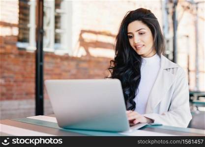 Adorable brunette woman in white clothes sitting outdoors at cafe using her laptop computer communicating with friends online using free internet connection. People, lifestyle, technology concept