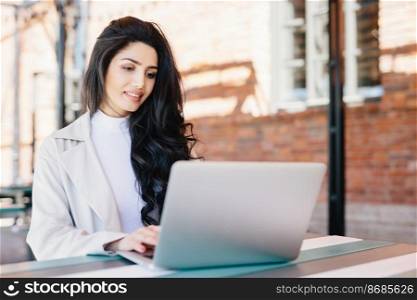Adorable brunette woman in white clothes sitting outdoors at cafe using her laptop computer communicating with friends online using free internet connection. People, lifestyle, technology concept