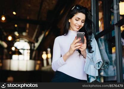 Adorable brunette woman with happy look having sunglasses on head and wearing white blouse siting over modern cafe interior, using cell phone, checking newsfeed on her social network accounts.