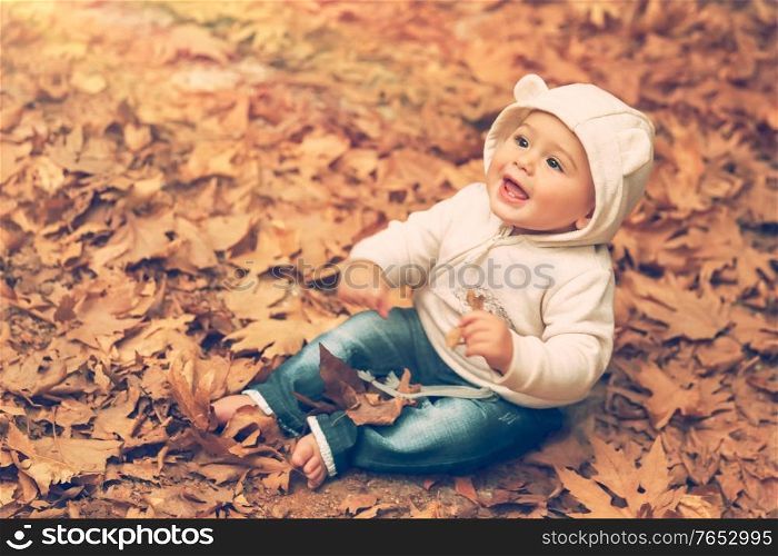 Adorable child sitting on the ground covered with dry tree leaves in warm autumn day and with wonder looking up to the sun, happy autumnal time