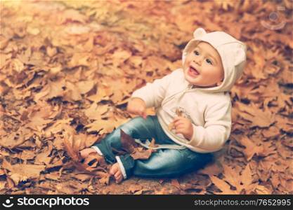 Adorable child sitting on the ground covered with dry tree leaves in warm autumn day and with wonder looking up to the sun, happy autumnal time