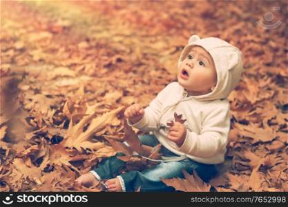 Adorable child sitting on the ground covered with dry tree leaves in warm autumn day and with wonder looking up to the sun lights, happy autumnal time