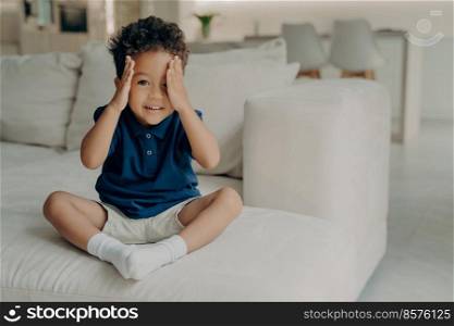 Adorable curly afro american boy in casual wear sitting on comfy big white couch and ready to play, stopping counting and uncovering his eyes, about to go searching in game of hide and seek. Cute little boy with curly hair enjoying game of hide and seek at home