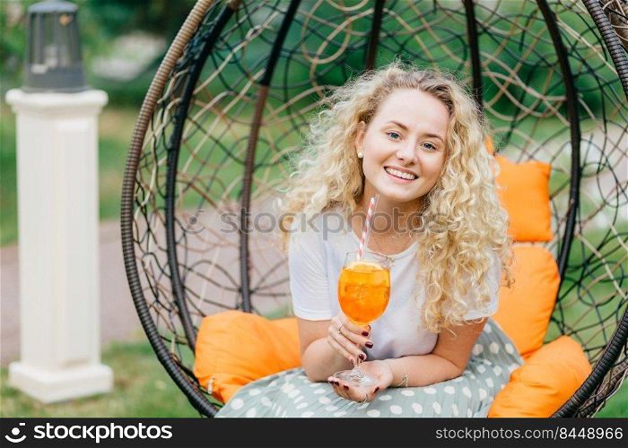 Adorable curly young female with curly bushy hair, holds glass of orange cocktail, poses in hanging outdoor chair, expresses positiveness, has pleasant talk with interlocutor. Leisure concept