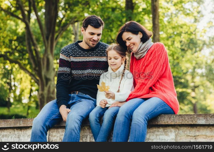 Adorable cute lovely small chld sits between her parents outdoors, holds leaf in hands, spend weekends together. Friendly young family enjoy togetherness, have happy expressions, sit on bridge