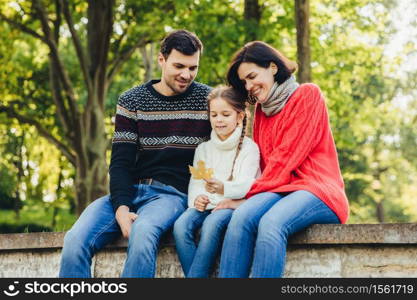 Adorable cute lovely small chld sits between her parents outdoors, holds leaf in hands, spend weekends together. Friendly young family enjoy togetherness, have happy expressions, sit on bridge