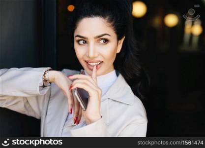Adorable dark-haired woman with appealing eyes, make-up, well-shaped lips and pure skin having red manicure holding her finger on white teeth holding smartphone in hand looking mysteriously aside