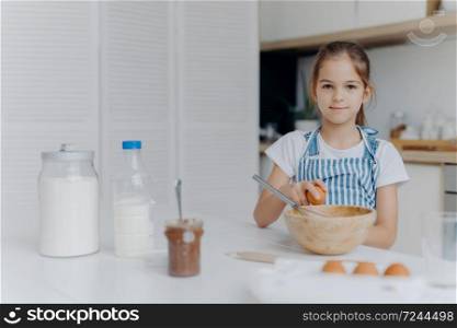 Adorable European child enjoys cooking activity, breaks egg in bowl, whisks ingredients, prepares tasty pastry, being young cook, wears apron, poses against kitchen interior, learns how to make cake