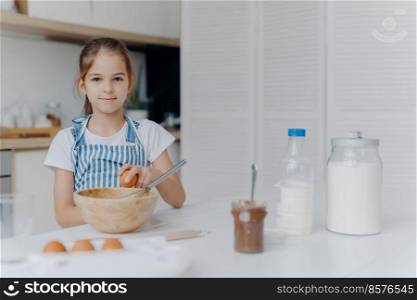 Adorable European child enjoys cooking activity, breaks egg in bowl, whisks ingredients, prepares tasty pastry, being young cook, wears apron, poses against kitchen interior, learns how to make cake