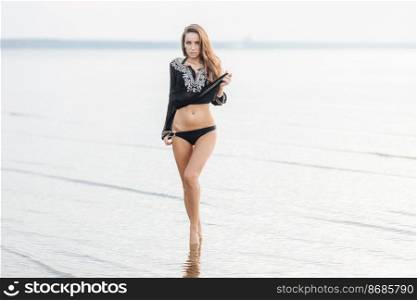 Adorable female in bikini demonstrates perfect body shape while stands at coastline, has slender legs, appealing appearance with make up. Relaxed attractive woman in swimsuit sunbathes outdoor