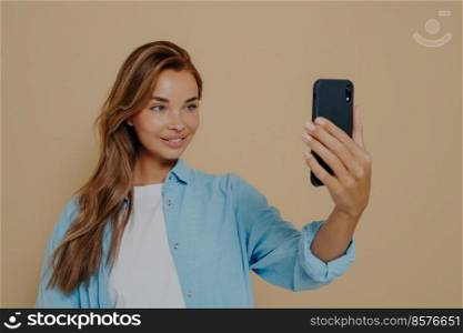 Adorable female model poses for making selfie, uses modern cell phone, dressed in blue oversized shirt over white tshirt, stands against pastel beige studio background, with happy facial expression. Adorable female model poses for making selfie in studio