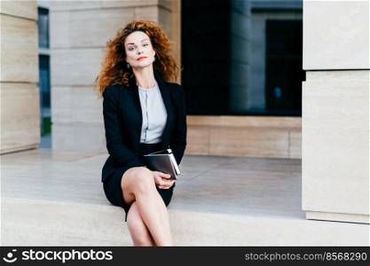 Adorable female model with bushy wavy hair, red lips and shining eyes, dressed formally while crossing her legs, holding tablet with pocket book, looking confidently into camera. Business concept
