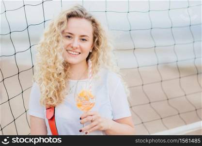 Adorable glad young European woman has bushy curly hair, smiles gently, holds glass of orange cocktail, stands near tennis net with copy space right for your advertisement or promotional text
