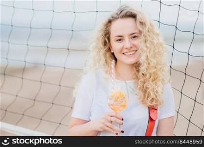 Adorable glad young European woman has bushy curly hair, smiles gently, holds glass of orange cocktail, stands near tennis net with copy space right for your advertisement or promotional text