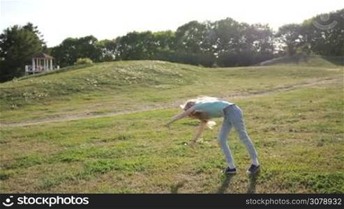 Adorable gymnastic girl with long blond hair doing her exercises on park lawn in glow of beautiful sunset. Sporty teenage girl doing cartwheel at green meadow against wonderful landscape. Slow motion. Steadicam stabilized shot.