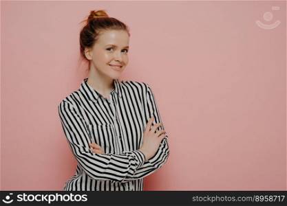 Adorable happy ginger teenage girl in striped shirt with hair in bun looking at camera with bright smile and expressing positive emotions, standing sideways with crossed arms against pink background. Positive young ginger lady with crossed arms smiling at camera