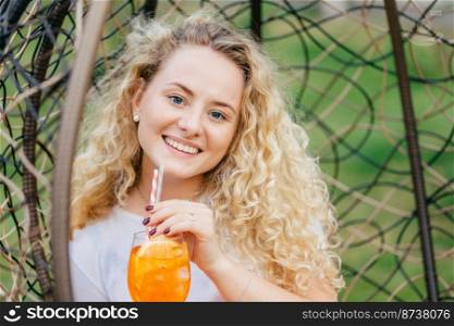 Adorable light haired positive female enjoys summer vacation, drinks fresh orange cocktail while sits in hanging chair, smiles positively, spends free time with boyfriend. People and recreation