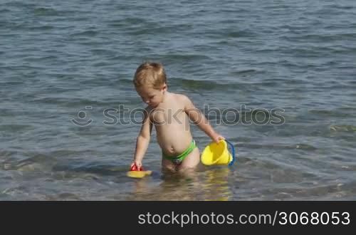 Adorable little boy playing in the sea enjoying the summer sunshine as he paddles in the water carrying his plastic toys