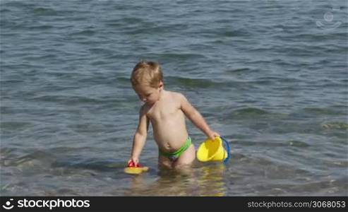 Adorable little boy playing in the sea enjoying the summer sunshine as he paddles in the water carrying his plastic toys