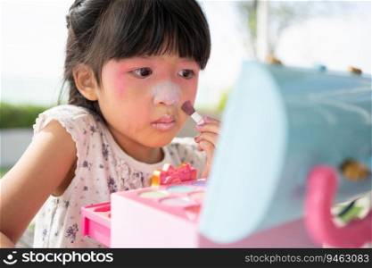 Adorable little child Asian girl paints her mouth with pink children heads and looks in the mirror. A child plays at home in a toy beauty salon. Increase learning development for preschoolers.