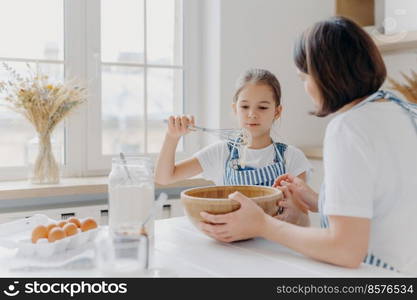 Adorable little child in apron shows whisk with white cream, wears apron, cooks together with mommy, pose in spacious kitchen together, make cake for special event. Children, help about house