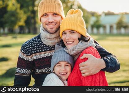 Adorable little child stand close to her affectionate parents, enjoy spending time together, embrace each other, smile happily, look directly into camera. Pleasant unforgettable moments with family