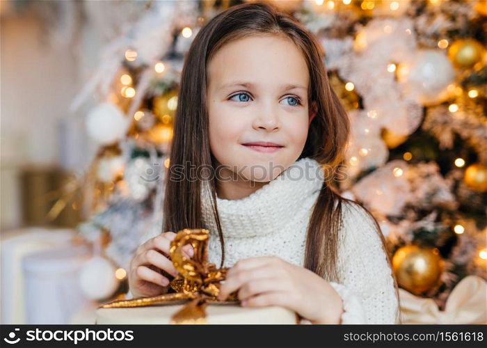 Adorable little female child with warm blue eyes, long dark hair, wears knitted warm white sweater, holds present stands against decorated New Year tree, looks aside on her affectionate parents