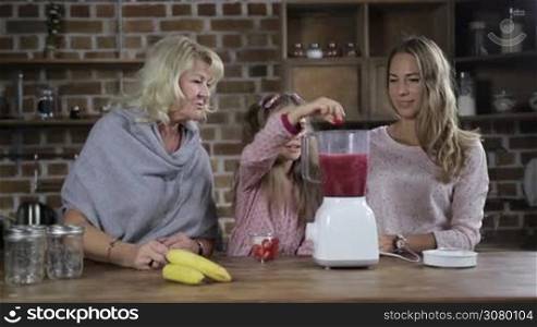 Adorable little girl adding strawberries into blender shaker jug helping her beautiful mother and grandmother to prepare detox raw fresh juice. Positive family making fresh berry smoothie together in the kitchen. Slow motion.
