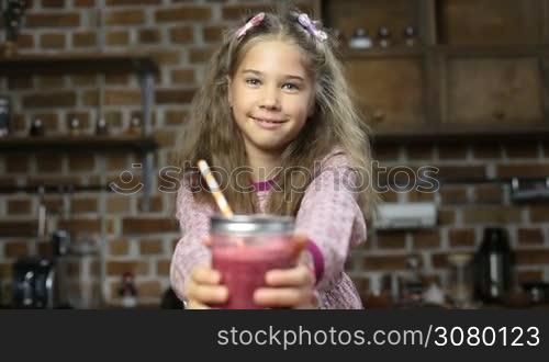 Adorable little girl offering delicious red berry smoothie in mason jar with drinking straw and smiling over domestic kitchen background. Lovely girl holding glass jar of fresh blended strawberry smoothie at home.