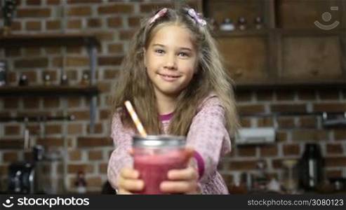 Adorable little girl offering delicious red berry smoothie in mason jar with drinking straw and smiling over domestic kitchen background. Lovely girl holding glass jar of fresh blended strawberry smoothie at home.