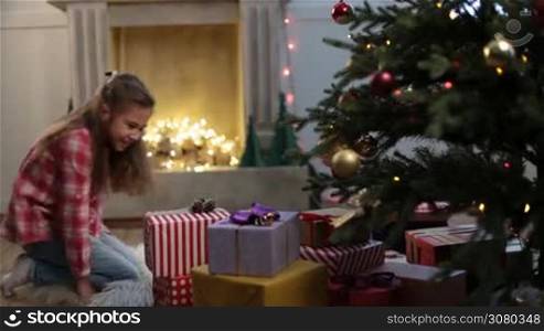 Adorable little girl runs to Christmas tree in the morning and starts looking for gifts. Excited cute daughter with long brown hair searching for her presents under xmas tree during winter holidays. Dolly shot.