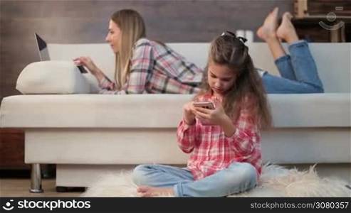 Adorable little girl sitting cross-legged on the floor wih smartphone while her beautiful mother lying on the couch and working on laptop at home. Daughter surfing social networks on mobile phone while her freelance mom using laptop pc. Dolly shot.