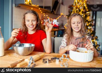 Adorable little girls baking cookies background of lights and Christmas tree. Happy kids celebrating holiday.. Little girls making Christmas gingerbread house at fireplace in decorated living room.