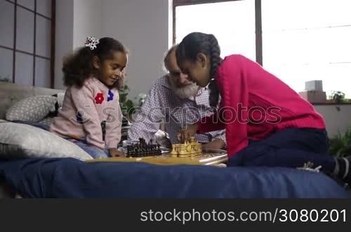 Adorable little sisters playing chess with help of handsome grandfather while sitting on bed at home. Pensive girl moving a pawn and her younger sister thinking intensely about the move. Caring grandpa giving advice to granddaughter about the game.