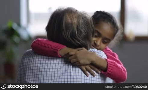 Adorable mixed race granddaughter embracing her caucasian grandpa, expressing love and tenderness over domestic interior. Portrait of smiling happy little girl hugging her beloved grandfathther at home.