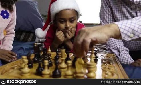 Adorable mixed race little girl in santa hat wathcing chess game between grandfather and her sister and smiling. Joyful cute child helping her younger sister to make a move while family playing chess board game on christmas eve. Dolly shot. Slo mo.
