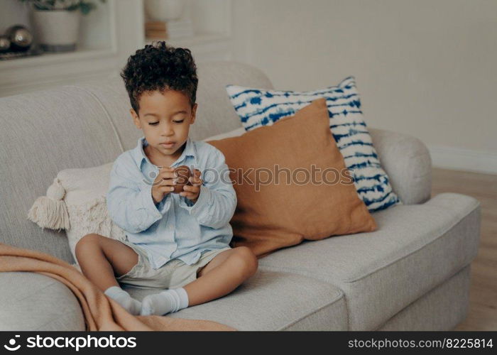 Adorable mulatto baby boy in casual clothes sitting on sofa decorated with pillows, holding chocolate egg and looking with interest expression on his face. Children and sweets concept. Adorable black baby boy in casual clothes with sweet chocolate egg
