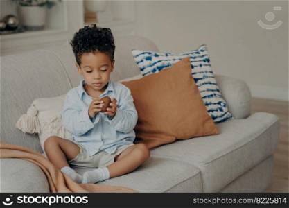 Adorable mulatto baby boy in casual clothes sitting on sofa decorated with pillows, holding chocolate egg and looking with interest expression on his face. Children and sweets concept. Adorable black baby boy in casual clothes with sweet chocolate egg