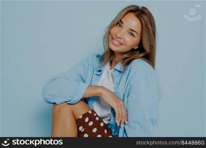 Adorable outgoing young woman with long hair and perfect healthy skin, smiling broadly at camera while sitting sideways, showing her straight white teeth. Beauty, style and fashion concept.. Studio photo of adorable outgoing young woman with long hair