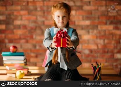 Adorable schoolgirl holds out a gift, front view. Female pupil sitting on the desk with stack of textbooks and apple on the top