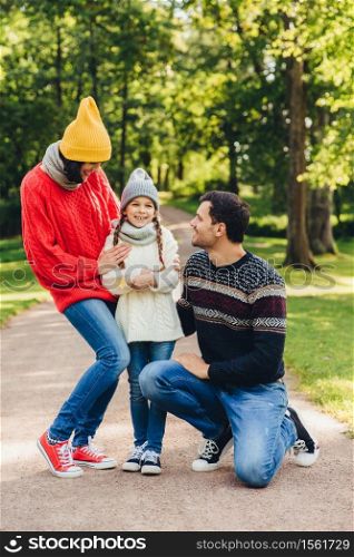 Adorable small cute child being glad to be supported by parents, shares her emotions after visiting kinder garten, has good relaxation in park. Model family walks in park, have happy expressions
