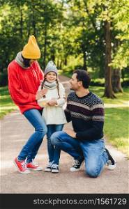 Adorable small cute child being glad to be supported by parents, shares her emotions after visiting kinder garten, has good relaxation in park. Model family walks in park, have happy expressions