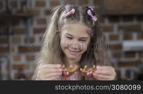 Adorable small girl fooling around with homemade cookies, using cookies as eyes over modern kitchen background. Smiling child holding christmas decorated reindeer pretzel cookies over her eyes and smiling at home.