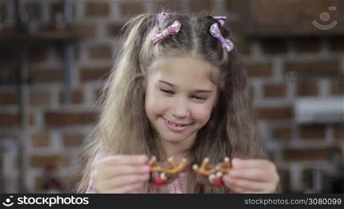 Adorable small girl fooling around with homemade cookies, using cookies as eyes over modern kitchen background. Smiling child holding christmas decorated reindeer pretzel cookies over her eyes and smiling at home.
