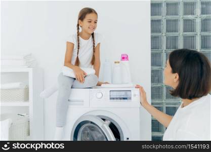 Adorable small girl with pigtails, poses on top of washer, holds white soft towel, looks gladfully at mother, talk about plans after washing. Brunette housewife loads washing machine, being busy